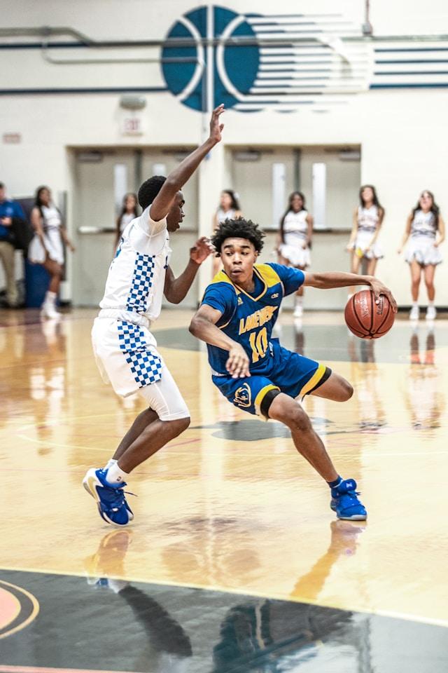 Two basketball players in action, with one dribbling the ball past the defender during a game while cheerleaders watch in the background